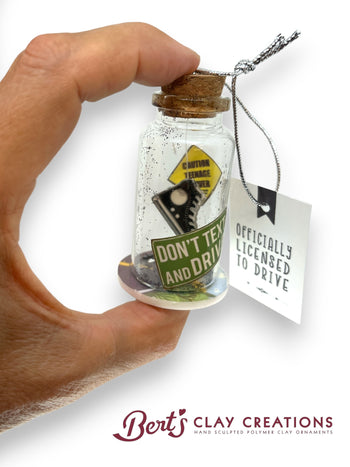 Hand holding a small glass bottle with a cork lid, containing a miniature sign and tag, with 'Bert's Clay Creations' branding.